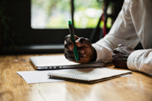 close up of hand holding a pen with a notepad and closed laptop nearby