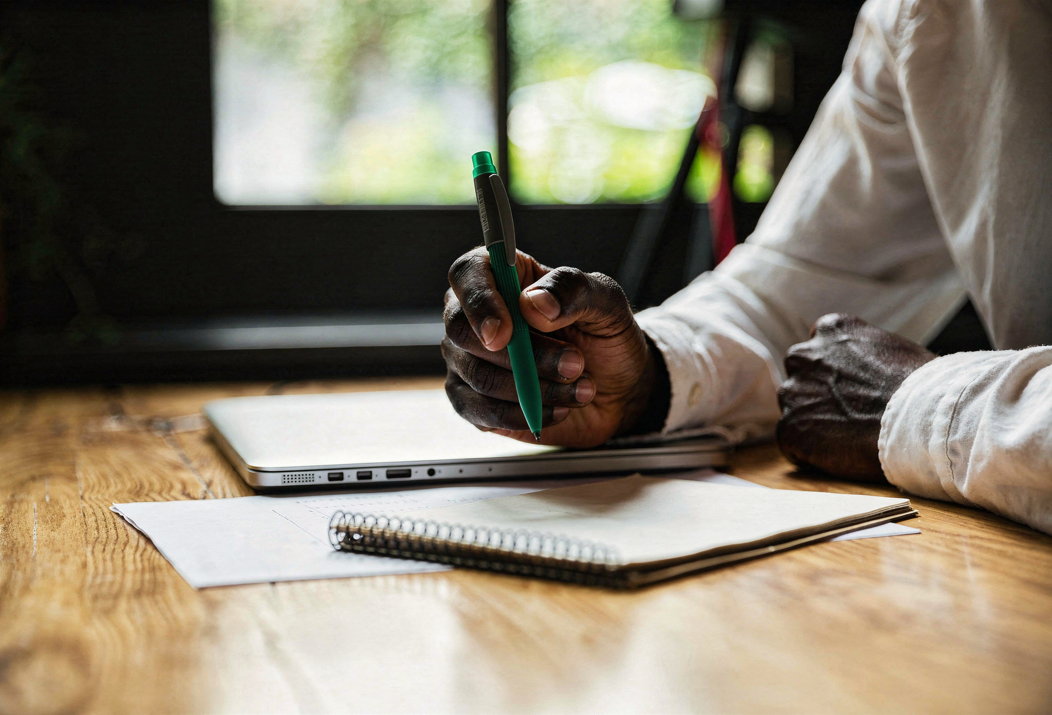 close up of hand holding a pen with a notepad and closed laptop nearby